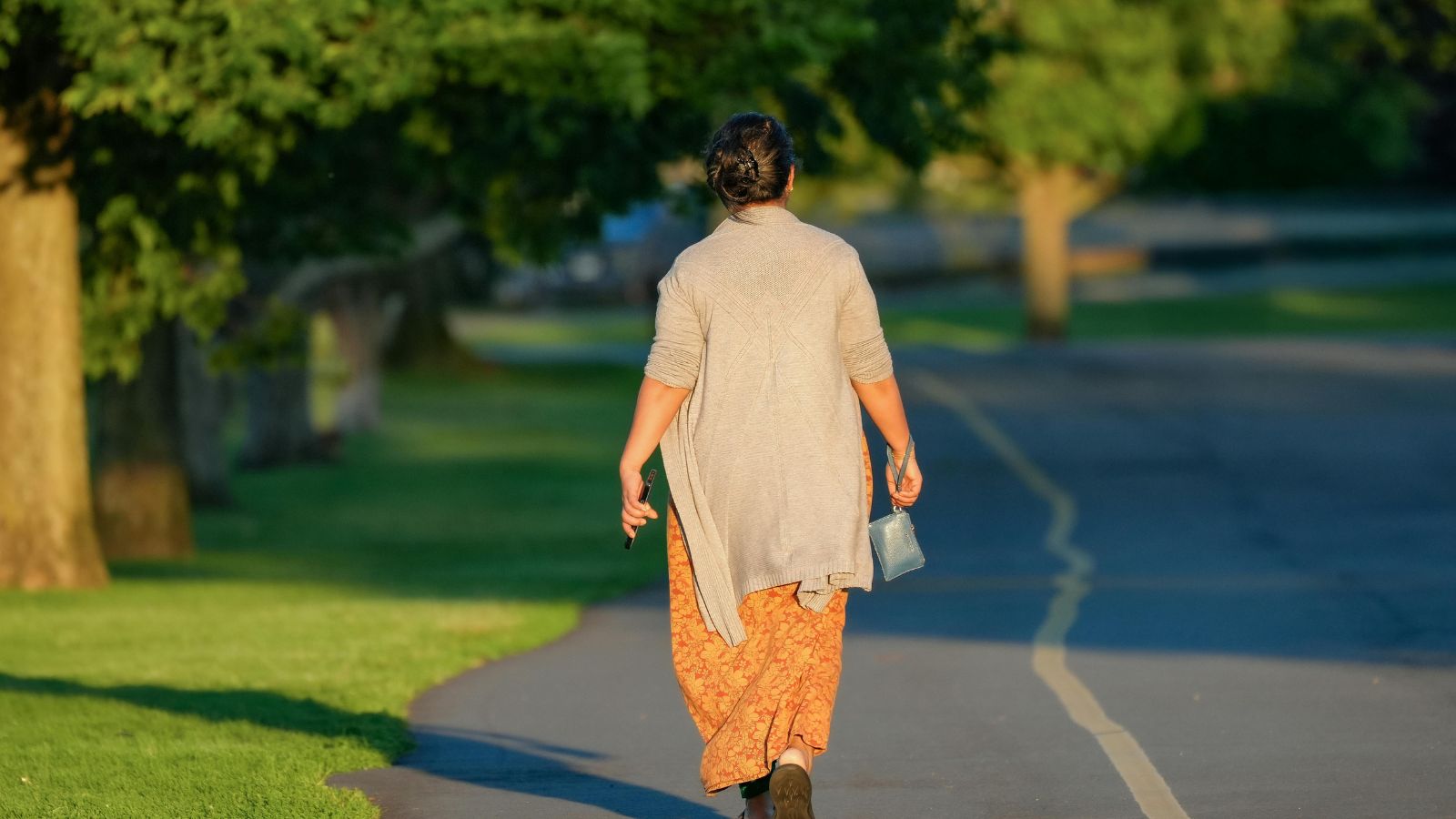 De dos, une femme marche seule sur une allée de parc dans la lumière de fin de journée.